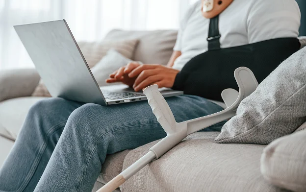 Hombre con el brazo inmovilizado usando su laptop, con muletas a un lado.