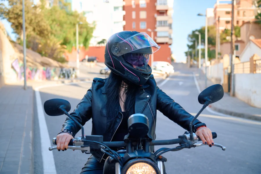 Motociclista con casco negro y chaqueta de cuero conduciendo una motocicleta por una calle urbana.